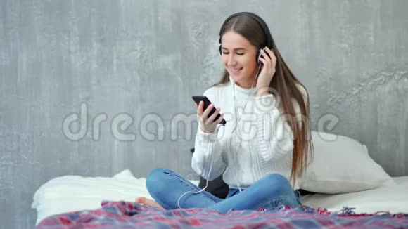快乐的少女微笑着坐在床上听着智能手机和耳机的音乐视频的预览图