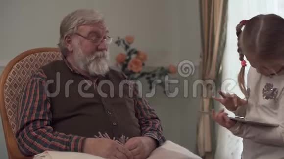 大胡子老人坐在大窗户附近的摇椅上看着他的孙女小女孩就在附近视频的预览图