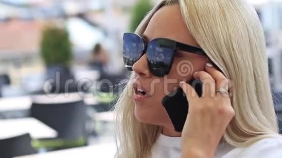 戴着太阳镜的年轻金发女郎在户外咖啡馆用手机近距离观看视频的预览图