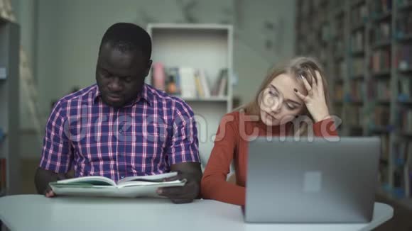 可爱疲惫的女孩带着笔记本电脑和她的非裔美国朋友在看书视频的预览图