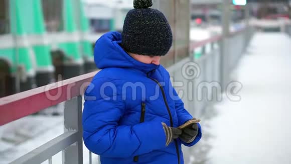 穿着蓝色羽绒服的男孩在街上读信息智能手机视频的预览图