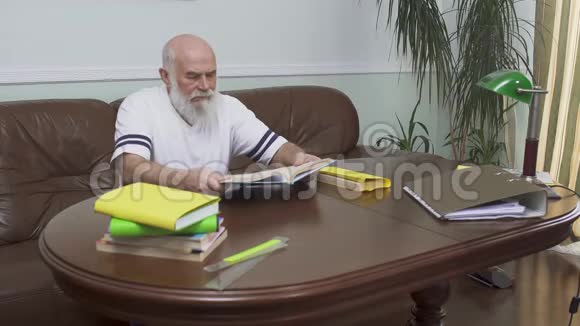 坐在木桌旁坐在皮革沙发上读着这本书白人老人享受视频的预览图