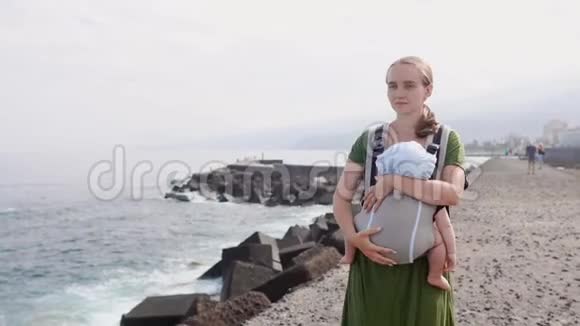 一位年轻的母亲正带着婴儿带着吊带在海滩上旅行视频的预览图
