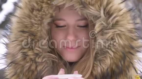 年轻的白种人女孩的特写照片她喝着一杯热饮料微笑着在冬天的街道上观看镜头视频的预览图