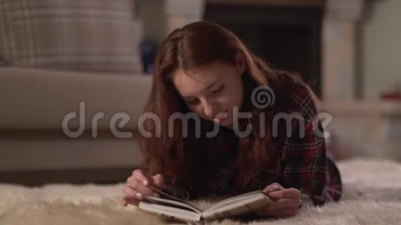 漂亮的年轻女人穿着格子衬衫躺在蓬松的地毯上近距离阅读这本书女士翻页微笑视频的预览图