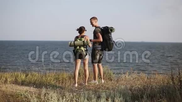 年轻夫妇站在山上尴尬地欣赏海景背包徒步旅行者背面视图视频的预览图