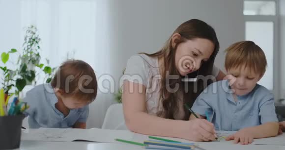 一位带着两个孩子在手机上说话的年轻母亲用铅笔画画并帮助孩子们用彩色画画视频的预览图