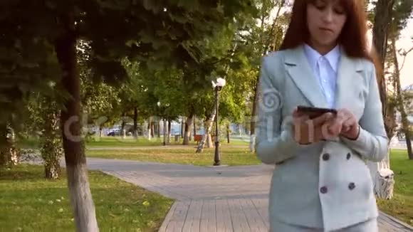 严肃的女商人走在公园的小巷里手里拿着平板电脑穿着轻便商务服的女士查看电子邮件视频的预览图