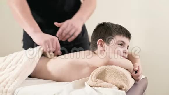 一位男性理疗师按摩师为躺在按摩床上的一个微笑的小男孩做一个疗愈放松的按摩护肩视频的预览图