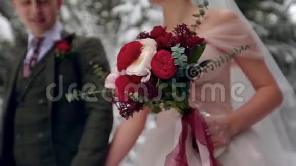 新娘携带一束由白色和红色玫瑰制成的花新婚夫妇手牵手走在雪林里视频的预览图