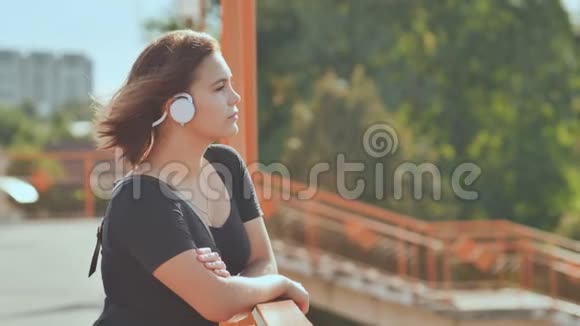 一个年轻的女孩戴着白色耳机在一个温暖的夏天晚上愉快地听音乐视频的预览图