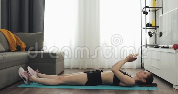 年轻的健身女性穿着运动运动服在使用智能手机上的秒表时从垫子上做运动她是视频的预览图