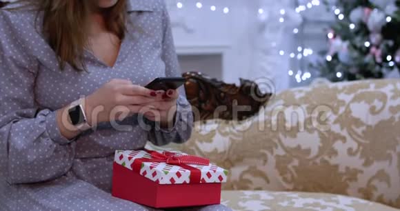 年轻的美女在圣诞夜用手机写短信坐在家里的沙发上视频的预览图