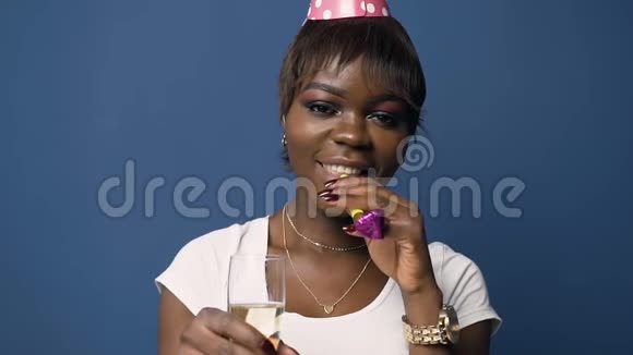 漂亮的非洲女人戴着生日帽拿着香槟杯在蓝色背景上吹着生日喇叭视频的预览图