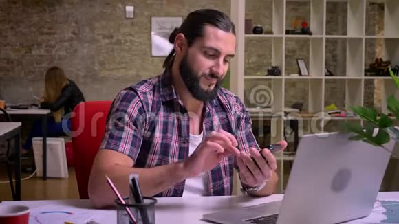 一个长着黑胡子留着长发极具创造力的考辛男性正在室内办公室里打电话视频的预览图