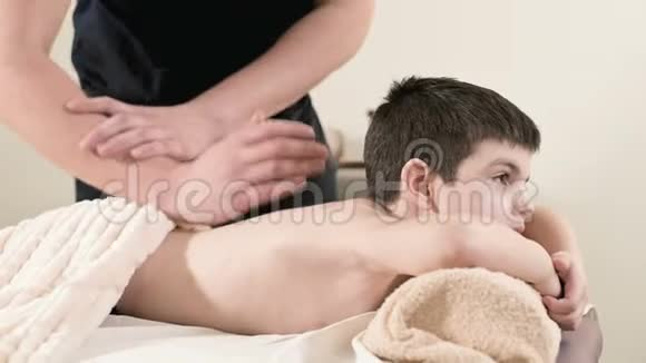 一位男性理疗师按摩师为躺在按摩床上的一个微笑的小男孩做一个疗愈放松的按摩护肩视频的预览图