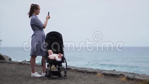 一位年轻的母亲站在海边一个婴儿坐在轮椅上在漂亮的智能手机上拍照视频的预览图
