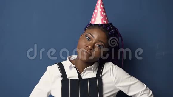 特写镜头英俊的非洲女孩戴着生日帽对着镜头微笑的蓝色背景视频的预览图