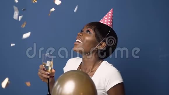快乐的非洲女孩戴着帽子一只手拿着香槟杯另一只手拿着气球看着飞舞的纸屑视频的预览图