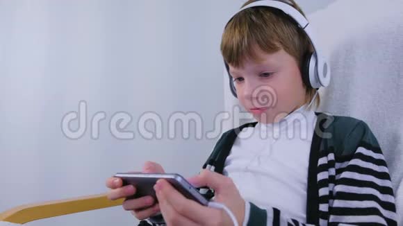 男孩正在玩一个耳机游戏智能手机坐在家里的扶手椅上视频的预览图