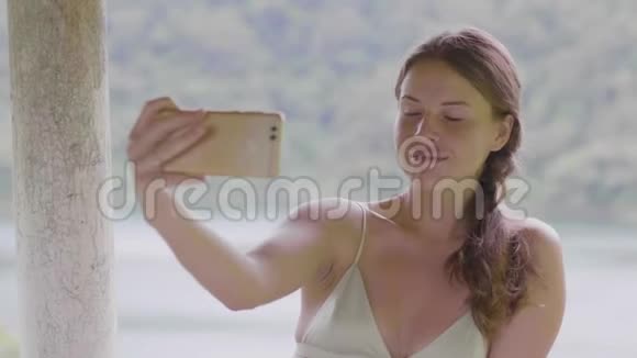 年轻女性使用手机拍摄湖泊和雨林景观的自拍照片旅行女孩用智能手机自拍视频的预览图