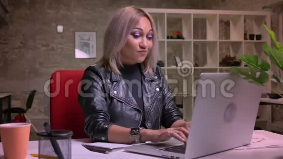 跳舞和放松的金发白种人女性她独自坐在电脑附近的砖砌办公室的桌面上视频的预览图