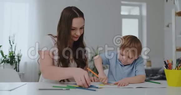 坐在客厅的桌子旁一位年轻的母亲教儿子拿铅笔教他画画视频的预览图