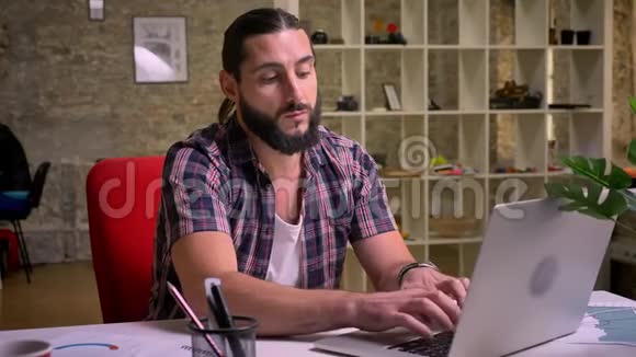 漂亮的白种人留着胡子的男人在工作场所坐着在笔记本电脑上打字时对手臂感到失望视频的预览图