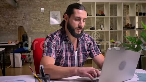 英俊的白种人工作的男性长着漂亮的胡须坐在他的桌面上停止他在电脑上的工作看着他视频的预览图