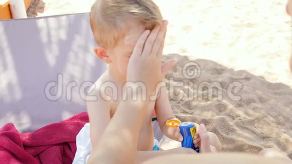 4k特写视频的年轻女子坐在沙滩上与蹒跚学步的儿子在日光浴妈妈给她涂防晒霜视频的预览图