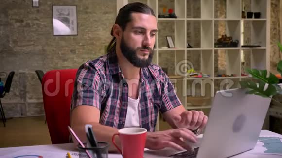 准确地看着白种人漂亮胡子男的屏幕随意的风格坐在灯光的现代办公室里工人视频的预览图