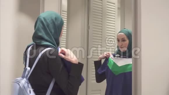 戴着头巾和背包的漂亮穆斯林女人在镜子前试穿衣服视频的预览图