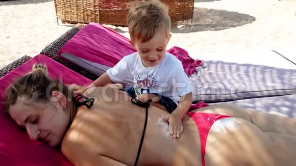 4k视频可爱的小男孩涂防紫外线乳液在母亲背上躺在海边的休息室视频的预览图
