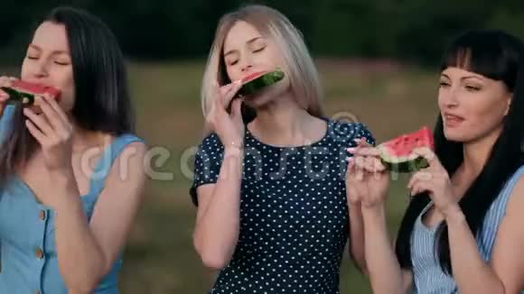 三个年轻迷人的女朋友在日落时穿着蓝色连衣裙吃西瓜和微笑视频的预览图