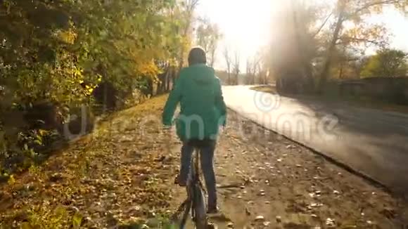 穿绿色夹克的年轻女孩骑自行车在阳光下在沥青路的一侧在秋天的公园里在一个背景下骑自行车视频的预览图