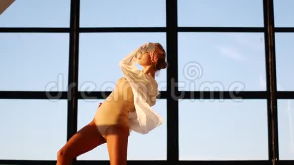 一个年轻而精力充沛的女孩跳着卑鄙的舞蹈在一个有大的全景窗户的阁楼工作室里一个穿着一身衬衫的女人视频的预览图