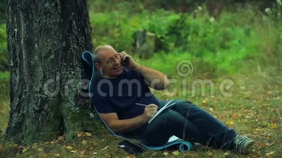 一个戴眼镜的微笑男人坐在公园的一棵树下用铅笔在笔记本上写字然后在电话上交谈视频的预览图