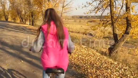 穿着粉红色夹克的女孩骑自行车双手握着方向盘在秋路上慢动作视频的预览图