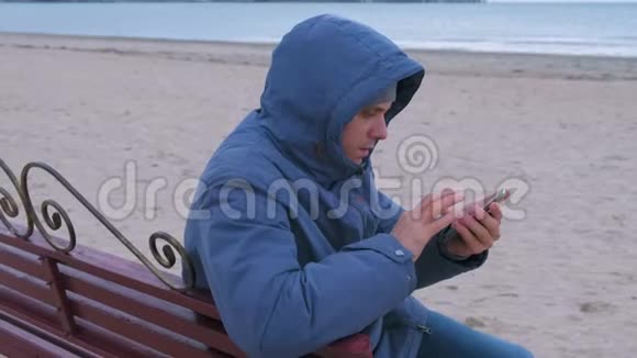 穿着蓝色羽绒服的男人坐在沙滩上的长凳上打着手机等待着答案视频的预览图