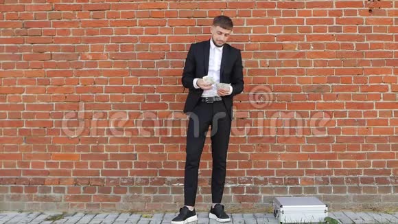一个穿着黑色西装的白种人年轻男子在基皮克墙的背景下清点钱财并将自己的一半投入其中视频的预览图
