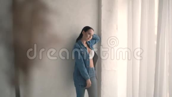 漂亮的女人穿着牛仔夹克和裤子在摄影棚窗户附近的镜头上摆姿势慢动作视频的预览图