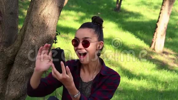 戴着太阳镜的漂亮女孩积极地笑着透过社交网络上的新闻坐在公园的长凳上视频的预览图