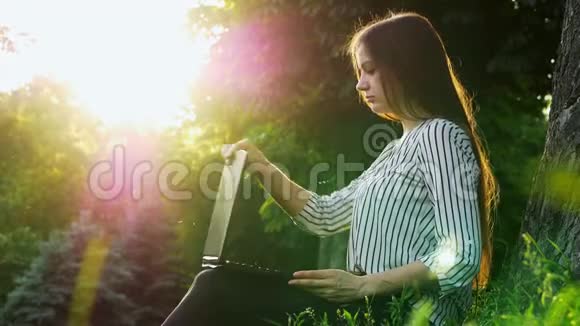 年轻漂亮的女商人在笔记本电脑上打字坐在靠近树的草地上女人在公园里用笔记本视频的预览图