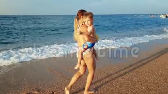 4k视频美丽的年轻女子双手抱着蹒跚学步的儿子在海滩上向他展示日落视频的预览图