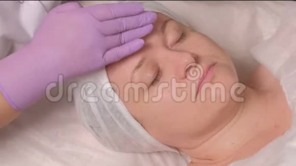 特写镜头戴着紫丁香手套的美容师用一种滋养凝胶涂在女性脸上一个中年妇女视频的预览图