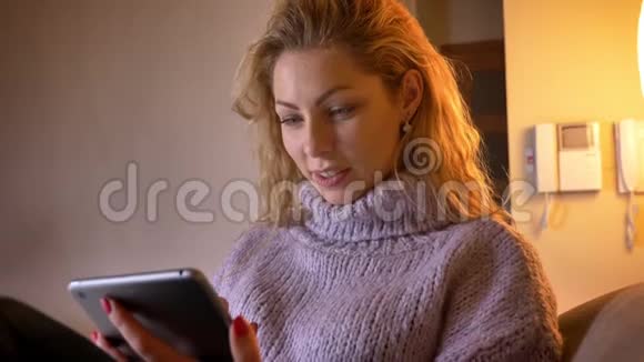 一位成年白种人金发女性在平板电脑上浏览并在舒适的公寓内欢快地笑着的特写镜头视频的预览图