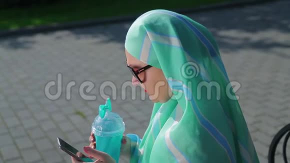 一位戴着浅色围巾的年轻穆斯林妇女坐在公园的长凳上喝着咖啡在送信者面前讲话视频的预览图