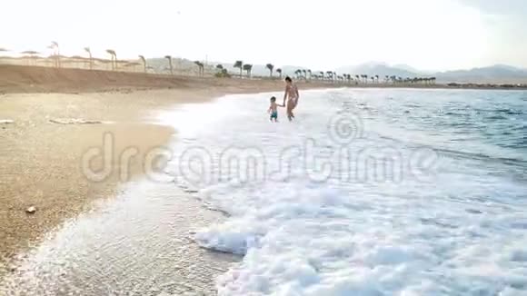 快乐的年轻母亲和她的小儿子在日落时在海浪中散步和跑步的视频家庭放松视频的预览图