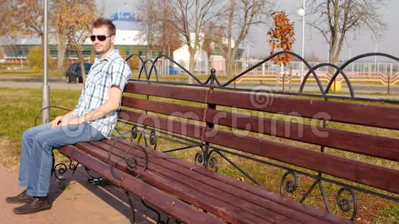 年轻英俊时尚的戴着黑眼镜的白种人坐在长凳上慢慢地看着镜头抄着视频的预览图