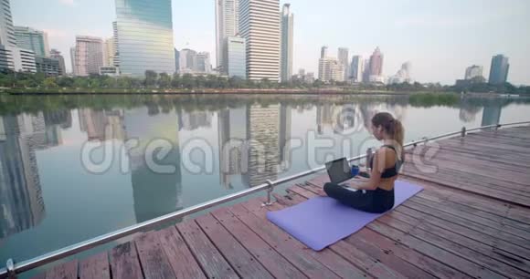 女人用手提电脑购买新的智能手机她坐在湖边的城市公园里与梅格波利斯市相对视频的预览图